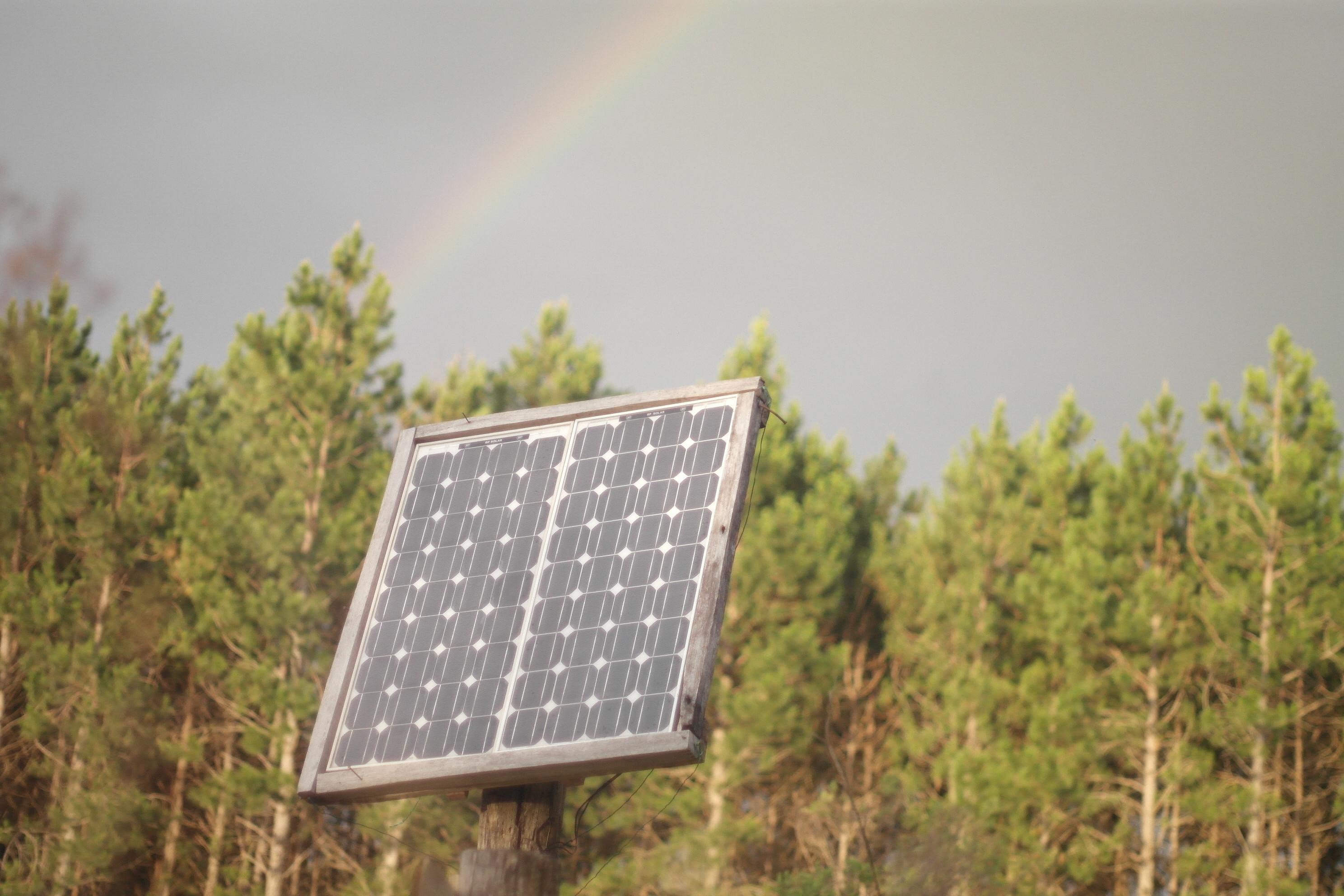 Imagen de dos placas solares de 100 wattios en un soporte hecho de piques sobre un poste.<br>En el fondo se puede apreciar un monte de pino y la parte de un arcoíris.